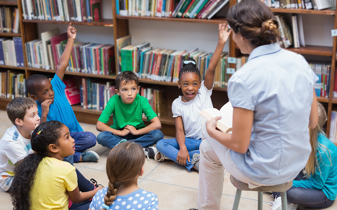 Students sit around teacher reading, some raising hands